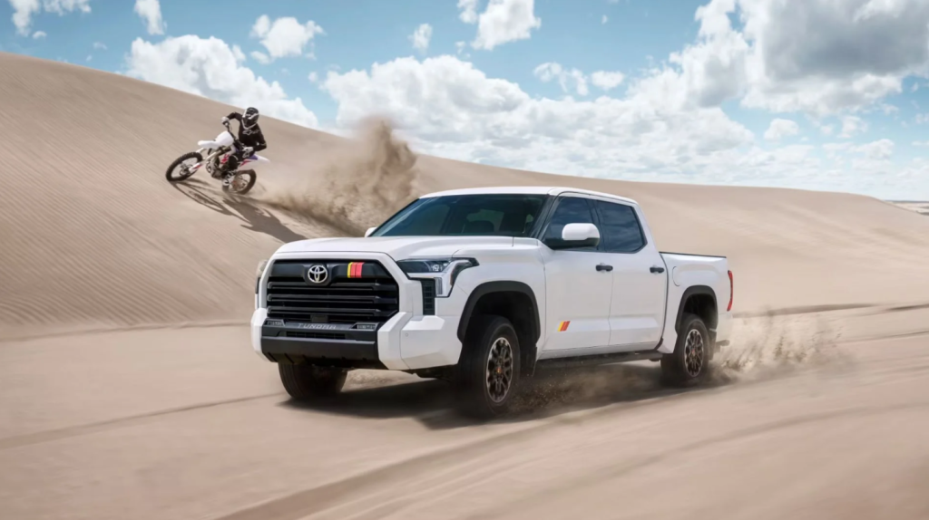 A man driving a white 2025 Toyota tundra through the sane with a dirt bike following closely behind.
