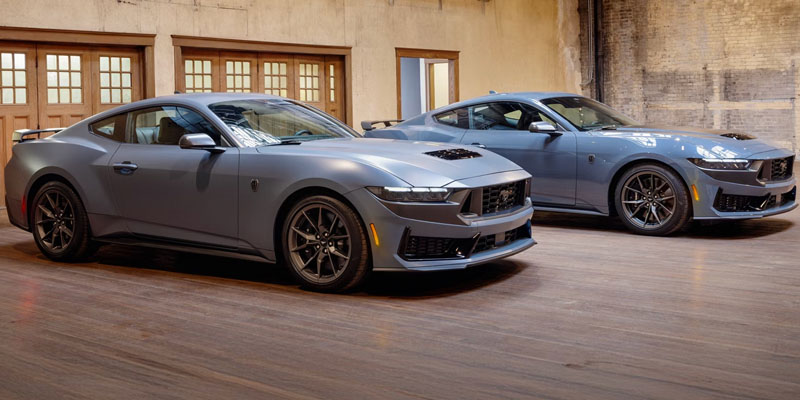 Two 2026 Ford Mustang models in a matte finish are parked side by side in an industrial-style interior, showcasing their sleek and aggressive design.